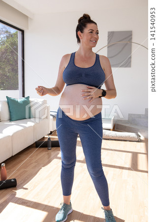 Pregnant woman placing hand on belly during living room workout with yoga mat and water bottles Pregnant woman placing hand on belly during living room workout with yoga mat and water bottles 141540953