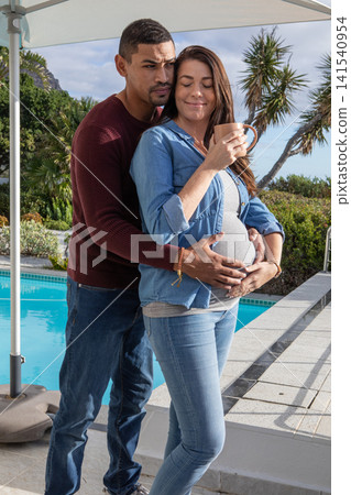 Diverse female couple hugging pregnant partner holding ceramic mug with patio umbrella by pool Diverse female couple hugging pregnant partner holding ceramic mug with patio umbrella by pool 141540954