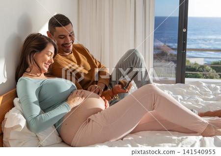 Diverse couple lying on bed with linens in bedroom, touching pregnant belly and holding tablet Diverse couple lying on bed with linens in bedroom, touching pregnant belly and holding tablet 141540955