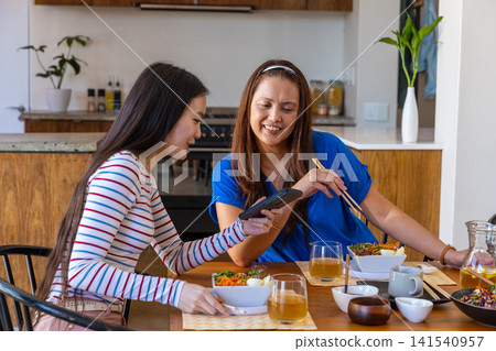 Asian mother and daughter sharing smartphone and using chopsticks to eat salad at table Asian mother and daughter sharing smartphone and using chopsticks to eat salad at table 141540957