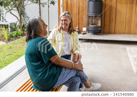 Diverse senior female friends chatting on patio bench holding mugs near stove, copy space 141540980