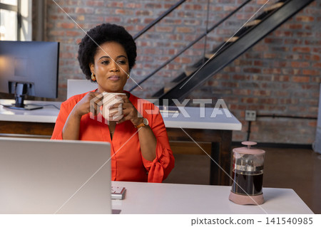 Mid adult african american woman holding coffee mug, working on laptop at desk in office Mid adult african american woman holding coffee mug, working on laptop at desk in office 141540985