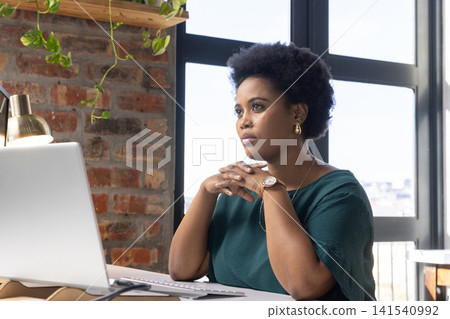 Mid thirties african american woman resting hands under chin at office desk with laptop Mid thirties african american woman resting hands under chin at office desk with laptop 141540992