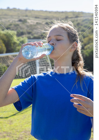 Teenage girl wearing blue athletic jersey lifting water bottle on soccer field goal net Teenage girl wearing blue athletic jersey lifting water bottle on soccer field goal net 141540994