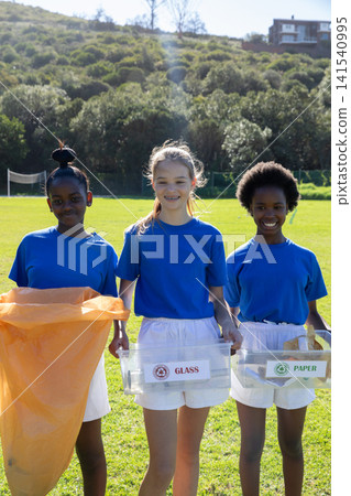 Diverse schoolgirls holding glass and paper bins and bag sorting recyclables on sports field Diverse schoolgirls holding glass and paper bins and bag sorting recyclables on sports field 141540995