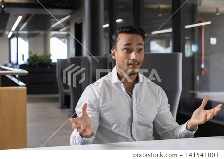 Indian man gesturing while speaking at white table in office with glass partitions 141541001