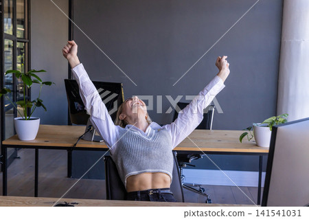 Woman sitting at modern office desk raising arms overhead with monitors Woman sitting at modern office desk raising arms overhead with monitors 141541031