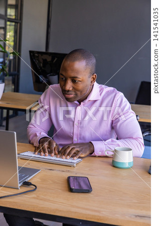 African american man typing on wireless keyboard in office with laptop, coffee mug African american man typing on wireless keyboard in office with laptop, coffee mug 141541035