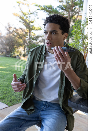 African american man sitting on stool at patio table holding smartphone close to mouth African american man sitting on stool at patio table holding smartphone close to mouth 141541045