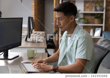 Asian man typing on wireless keyboard at open-plan office desk with laptop, mug, copy space Asian man typing on wireless keyboard at open-plan office desk with laptop, mug, copy space 141541048