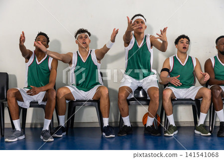 Male teammates in basketball uniforms shouting while sitting on chairs at court with basketball Male teammates in basketball uniforms shouting while sitting on chairs at court with basketball 141541068