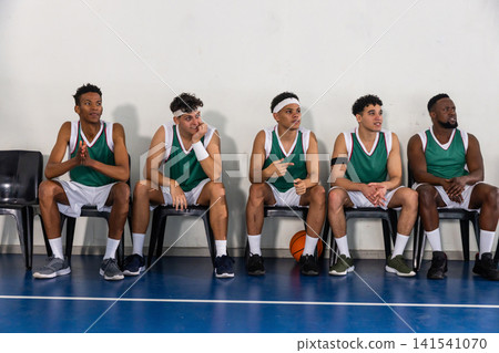 Diverse male basketball teammates resting on chairs wearing green uniforms with basketball Diverse male basketball teammates resting on chairs wearing green uniforms with basketball 141541070