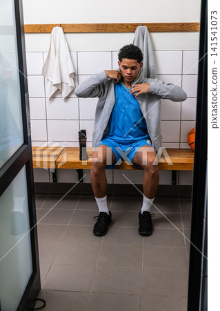 Teenage african american basketball player adjusting grey hoodie on bench in locker room Teenage african american basketball player adjusting grey hoodie on bench in locker room 141541073