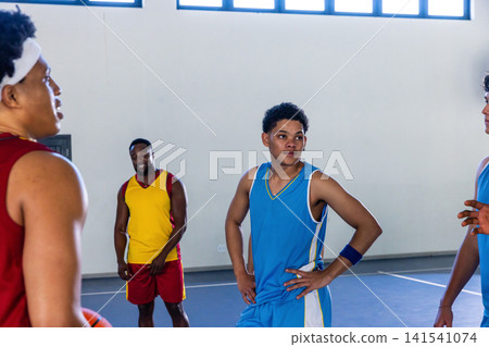 African american teen in blue jersey talking with teammates on court holding basketball African american teen in blue jersey talking with teammates on court holding basketball 141541074