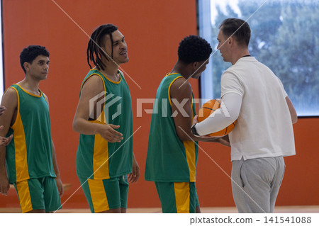 Diverse male basketball team in uniforms standing on court, coach handling basketball Diverse male basketball team in uniforms standing on court, coach handling basketball 141541088