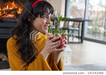 Woman wearing mustard sweater and red headband holding mug near fireplace in living room Woman wearing mustard sweater and red headband holding mug near fireplace in living room 141541098