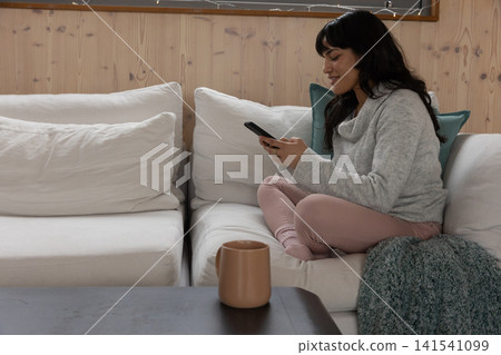 Woman sitting cross-legged on couch in living room with throw holding smartphone, copy space Woman sitting cross-legged on couch in living room with throw holding smartphone, copy space 141541099