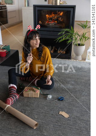 Woman in her thirties wrapping gifts using kraft paper sitting on rug by fireplace 141541101