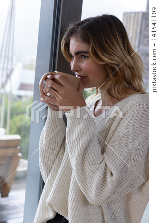 Woman standing beside glass sliding door holding ceramic mug at home in knit sweater, copy space Woman standing beside glass sliding door holding ceramic mug at home in knit sweater, copy space 141541109