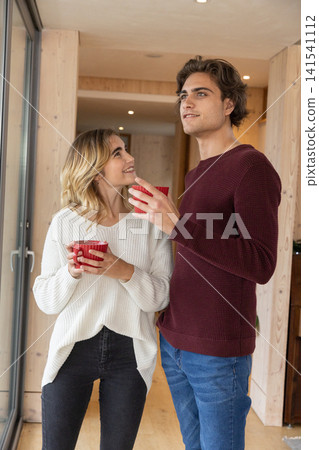 Couple wearing sweaters standing holding red mugs by sliding glass door in wood-paneled home Couple wearing sweaters standing holding red mugs by sliding glass door in wood-paneled home 141541112