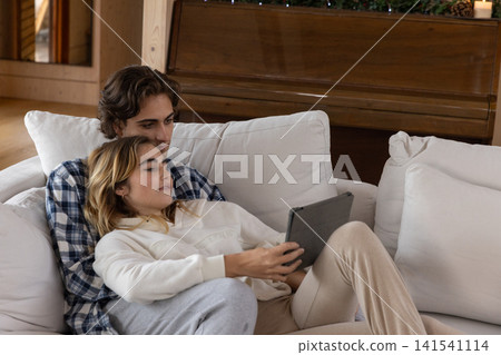 Couple cuddling on sofa in wood paneled room holding tablet near pine cones, candles on piano Couple cuddling on sofa in wood paneled room holding tablet near pine cones, candles on piano 141541114
