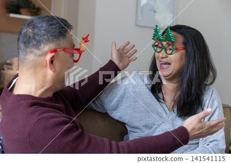 Asian couple sitting on brown couch in living room wearing novelty christmas glasses, laughing Asian couple sitting on brown couch in living room wearing novelty christmas glasses, laughing 141541115