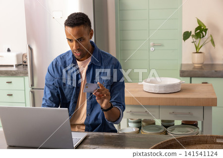 Mid adult african american man typing on laptop while holding credit card at home kitchen island Mid adult african american man typing on laptop while holding credit card at home kitchen island 141541124