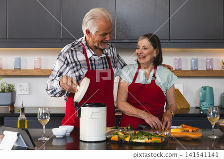 Senior couple cooking at kitchen countertop, chopping vegetables and holding lidded container Senior couple cooking at kitchen countertop, chopping vegetables and holding lidded container 141541141