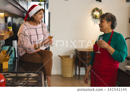 Diverse female friends conversing and holding wine glasses in kitchen in Santa hat and red apron Diverse female friends conversing and holding wine glasses in kitchen in Santa hat and red apron 141541145