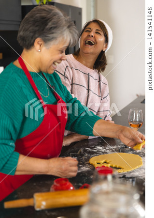 Senior mother and daughter rolling dough with red cutters at kitchen counter near wine glass 141541148