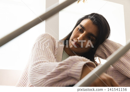 Asian woman wearing cardigan leaning on metal stair railing at home, gazing downward near window Asian woman wearing cardigan leaning on metal stair railing at home, gazing downward near window 141541156