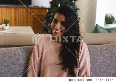 Asian woman sitting on gray sofa with green cushion at home by decorated Christmas tree 141541157