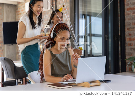 Diverse female coworkers approaching Hispanic woman using laptop with headset and cake at office Diverse female coworkers approaching Hispanic woman using laptop with headset and cake at office 141541187