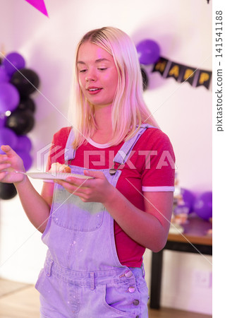 Female teen eating cake slice on white plate in home party room with balloons, banner Female teen eating cake slice on white plate in home party room with balloons, banner 141541188