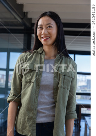 Asian woman standing smiling looking up in office with city view wearing green shirt and trousers Asian woman standing smiling looking up in office with city view wearing green shirt and trousers 141541189