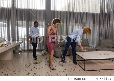 Diverse friends cleaning up confetti with broom around coffee table, wine glasses in living room Diverse friends cleaning up confetti with broom around coffee table, wine glasses in living room 141541195