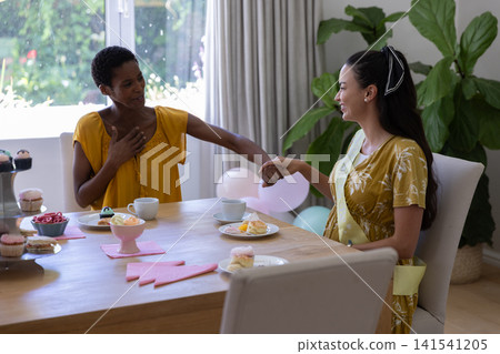 Diverse female friends holding hands across wooden dining table at home with pastries and balloons Diverse female friends holding hands across wooden dining table at home with pastries and balloons 141541205