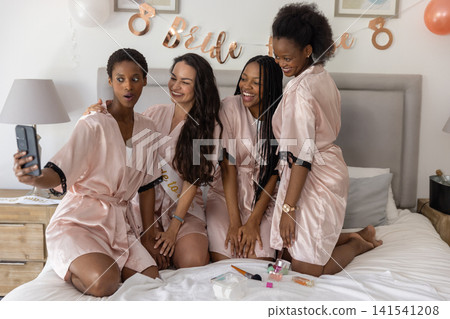 Diverse female friends taking smartphone selfie on bed wearing pink robes with Bride to Be sash Diverse female friends taking smartphone selfie on bed wearing pink robes with Bride to Be sash 141541208