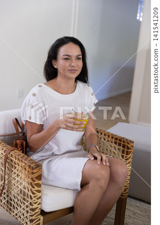 Woman sitting on woven rattan chair wearing white dress holding glass of orange juice at home Woman sitting on woven rattan chair wearing white dress holding glass of orange juice at home 141541209