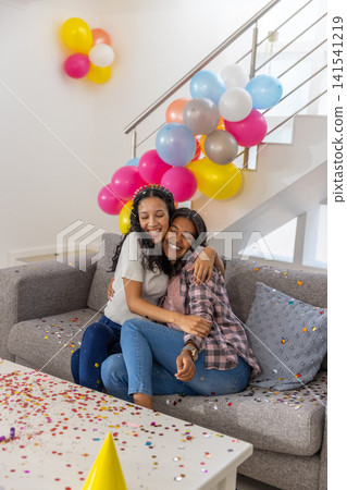 Diverse mom and teenage daughter hugging on grey sofa in living room with confetti, balloons Diverse mom and teenage daughter hugging on grey sofa in living room with confetti, balloons 141541219