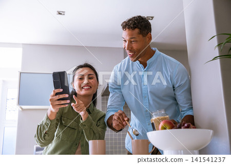 Diverse couple pointing at smartphone, scooping sugar from glass jar into mugs in kitchen Diverse couple pointing at smartphone, scooping sugar from glass jar into mugs in kitchen 141541237