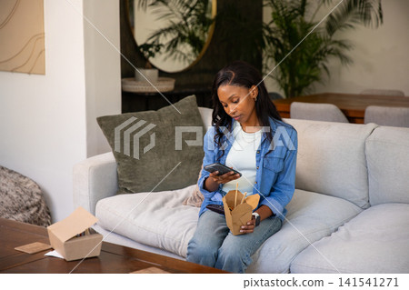 African American woman sitting on sofa in living room, holding smartphone, chopsticks and takeout African American woman sitting on sofa in living room, holding smartphone, chopsticks and takeout 141541271