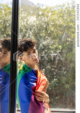 Man standing at window wearing rainbow pride flag and watching garden through glass pane Man standing at window wearing rainbow pride flag and watching garden through glass pane 141541842