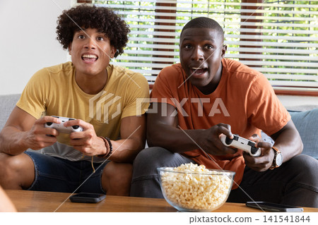 Diverse male friends gripping gamepads on sofa, smartphone and popcorn on table under blinds Diverse male friends gripping gamepads on sofa, smartphone and popcorn on table under blinds 141541844