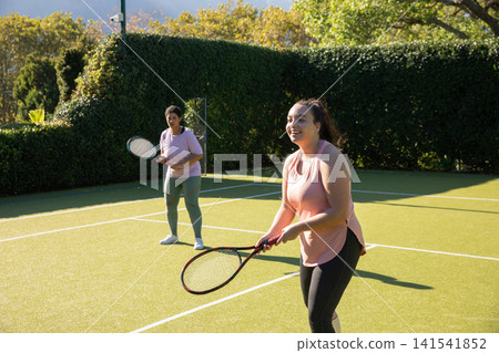 Diverse female friends practicing tennis on hedge-lined grass court and holding rackets, copy space Diverse female friends practicing tennis on hedge-lined grass court and holding rackets, copy space 141541852
