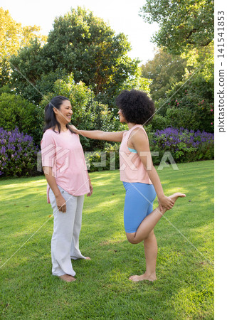 Mother guiding daughter stretching quads on lawn with purple shrubs in backyard Mother guiding daughter stretching quads on lawn with purple shrubs in backyard 141541853