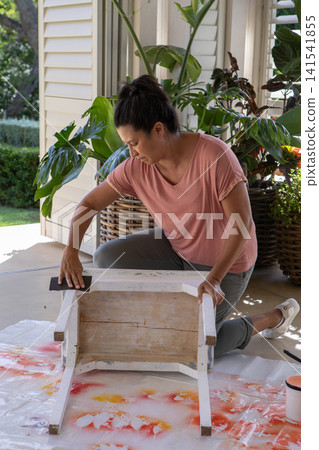 Hispanic woman kneeling on porch drop cloth sanding stool with sanding block next to paint can 141541855