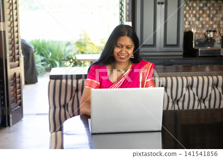 Mature indian woman wearing bright pink gold-striped sari sitting at home using laptop Mature indian woman wearing bright pink gold-striped sari sitting at home using laptop 141541856