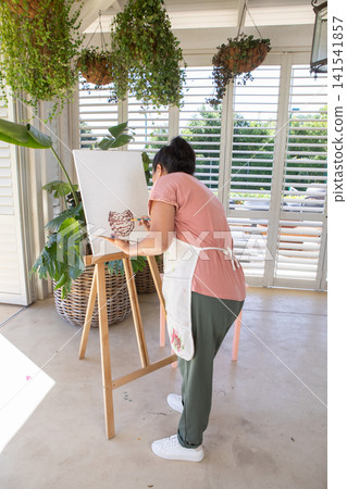 Woman in floral apron painting ceramic pitcher design on canvas at sunlit studio with potted plant Woman in floral apron painting ceramic pitcher design on canvas at sunlit studio with potted plant 141541857