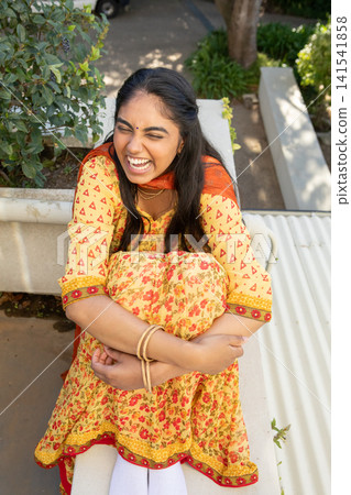Indian woman hugging knees while laughing on stone bench at garden terrace with red dupatta Indian woman hugging knees while laughing on stone bench at garden terrace with red dupatta 141541858